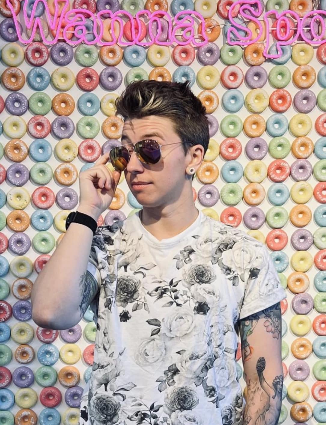 A picture of me, Rowan Bell. I have pale pink skin, short brown hair, and multiple tattoos on my arms. I'm wearing a white T-shirt with black and grey flowers, and I'm adjusting my sunglasses. I'm standing against a background image of multi-colored round cereal pieces.